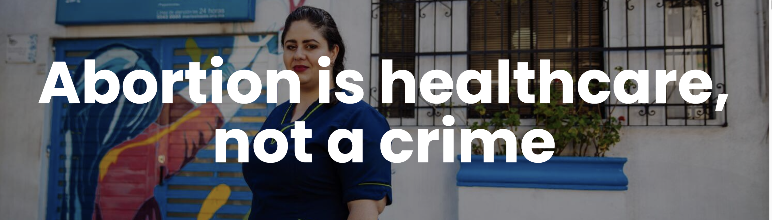 white text: abortion is healthcare not a crime, over a picture of a woman in a blue short sleeve shirt standing in front of railings and mural in a street